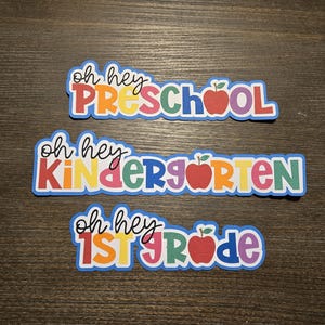 Puede incluir: Tres pancartas coloridas con el texto "Oh hey Preschool", "Oh hey Kindergarten" y "Oh hey 1st Grade". Cada pancarta tiene un gráfico de manzana roja.