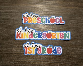 Puede incluir: Tres pancartas coloridas con el texto "Oh hey Preschool", "Oh hey Kindergarten" y "Oh hey 1st Grade". Cada pancarta tiene un gráfico de manzana roja.