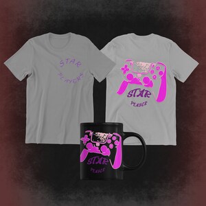 Puede incluir: Dos camisetas grises con un gráfico de mando de videojuego rosa y el texto "STAR PLAYER". Una camiseta tiene el texto "STAR PLAYERS" en un círculo. Una taza negra con el mismo gráfico de mando de videojuego rosa y el texto "STAR PLAYER".