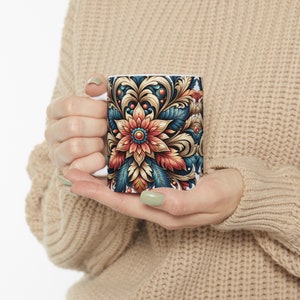 Puede incluir: Una taza de cerámica blanca con un diseño floral colorido. El diseño presenta una flor roja grande con un centro dorado rodeado de intrincados patrones dorados y azules.