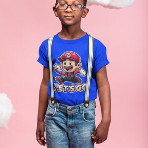 May include: A young person wearing a blue t-shirt with a cartoon character and the text "LET'S GO" printed on it. The shirt has white suspenders and the person is wearing blue jeans.