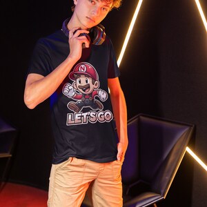 May include: A young person wearing a black t-shirt with a cartoon character and the text "LET'S GO". The character is wearing a red hat with a letter "N" on it. The person is wearing khaki pants and has curly brown hair.