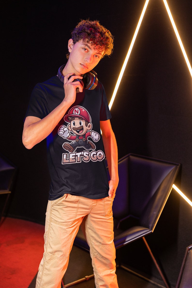 May include: A young person wearing a black t-shirt with a cartoon character and the text "LET'S GO". The character is wearing a red hat with a letter "N" on it. The person is wearing khaki pants and has curly brown hair.