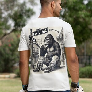 Puede incluir: Camiseta blanca con una ilustración en blanco y negro de un gorila sentado en una esquina de la calle. El letrero de la calle dice "LA LISA".