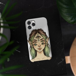 Op de afbeelding: Een witte telefoonhoes met een kleurrijke illustratie van een persoon met meerdere ogen en elfenoren. De persoon heeft groen haar en draagt ​​een zilveren neusring.