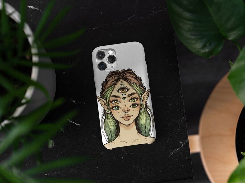 Op de afbeelding: Een witte telefoonhoes met een kleurrijke illustratie van een persoon met meerdere ogen en elfenoren. De persoon heeft groen haar en draagt ​​een zilveren neusring.