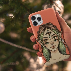 Op de afbeelding: Een koraalroze telefoonhoesje met een kleurrijke illustratie van een vrouw met meerdere ogen en elfenoren.
