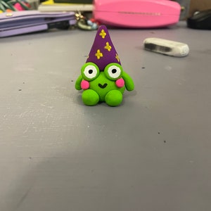 Puede incluir: Una rana verde con un sombrero morado con estrellas amarillas. La rana tiene mejillas rosadas y una expresión feliz.