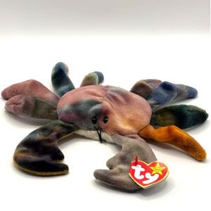 May include: A plush crab toy with a tie-dye pattern in shades of pink, blue, green, and orange. The crab has black eyes and a red and yellow heart-shaped tag attached to one of its claws. The toy is designed to be soft and cuddly.