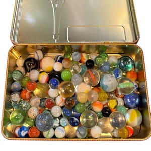May include: A collection of colorful glass marbles in a vintage metal tin. The marbles vary in size, color, and design, including swirls, solid colors, and transparent styles. The tin is open, revealing the assortment of marbles.
