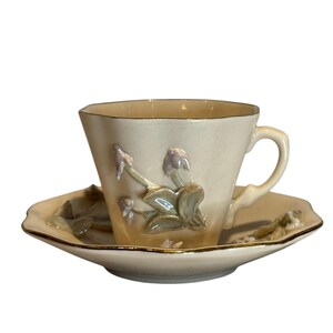Peut inclure: Une tasse à thé et une soucoupe vintage avec un motif floral. La tasse et la soucoupe sont de couleur crème avec une bordure dorée. Le motif floral présente des fleurs et des feuilles délicates dans des tons de vert, rose et violet.