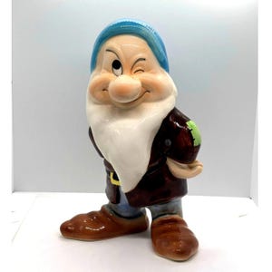 Op de afbeelding: Een keramieken beeldje van een knipogende dwerg met een lange witte baard, gekleed in een blauwe muts, een bruin jasje met een groene patch op de schouder, een blauwe broek en bruine schoenen. Het figuurtje heeft een grote neus en een vrolijke blik.