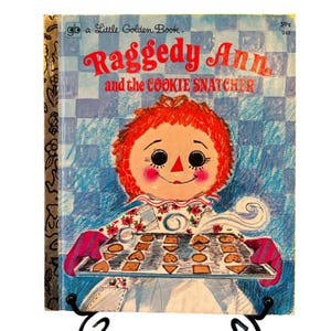 May include: A vintage Little Golden Book titled "Raggedy Ann and the Cookie Snatcher." The cover features a smiling Raggedy Ann character with red hair, holding a tray of cookies. The background is a blue and white checkered pattern.