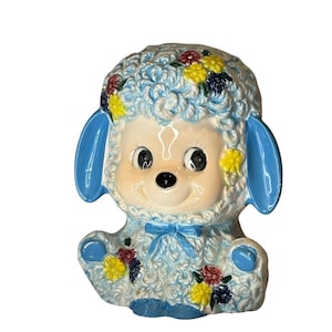 May include: A blue ceramic lamb piggy bank with a fluffy texture and floral details. The lamb has a smiling face with large eyes and a blue bow.
