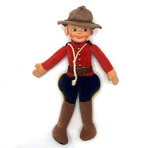 Könnte beinhalten: Eine Puppe, die als Offizier der Royal Canadian Mounted Police gekleidet ist, mit einem braunen Filzhut, einer roten langärmeligen Jacke mit goldenen Knöpfen, einem braunen Gürtel, dunkelblauen Reithosen mit gelben Akzenten und braunen Stiefeln.