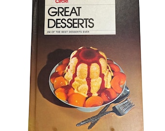 Może przedstawiać: Antyczna książka kucharska zatytułowana "Great Desserts" autorstwa Family Circle, przedstawiająca zdjęcie żółtego deseru polanego czerwonym sosem i otoczonego plastrami brzoskwiń na białym talerzu.