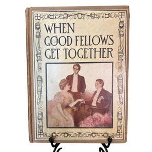 May include: Antique book titled "When Good Fellows Get Together." The cover features a sepia-toned illustration of three people, with the title in bold, black letters. The book is displayed on a black metal stand.