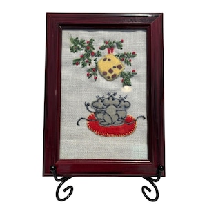 May include: Framed embroidery art featuring a whimsical design. The artwork depicts two gray mice on a red cushion beneath a sprig of berries and a yellow cheese wedge. The frame is dark red, and the piece is displayed on a black metal stand.
