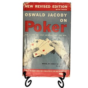 Può includere: Una copertina di libro vintage con un titolo rosso e bianco "Oswald Jacoby on Poker" con un'immagine di mani che tengono carte da gioco. Il testo sulla copertina recita "Come giocare al grande gioco americano - e vincere!" e "Insolito, ma corretto questa volta. Vedi pagina 4."