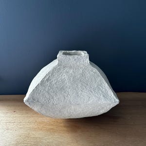 May include: A white ceramic vase with a textured surface. The vase has a wide, rounded base and a narrow opening at the top.