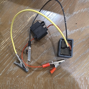 Puede incluir: Un conjunto de componentes eléctricos que presenta una carcasa rectangular negra con un cable amarillo y un cable negro, conectado a un portafusibles negro y dos pinzas de cocodrilo metálicas con fundas protectoras rojas y plateadas.