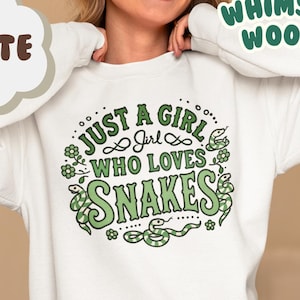 Puede incluir: Sudadera blanca con texto verde que dice "Just a girl who loves snakes". El texto está rodeado de ilustraciones de serpientes y flores verdes.
