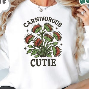 May include: White sweatshirt with a graphic of a Venus flytrap plant. The text on the shirt reads "CARNIVOROUS CUTIE" in a whimsical font. The design features green and red colors. The brand name "Whimsy's Woods" is also visible.