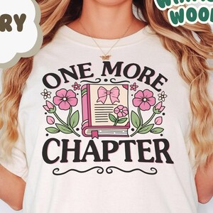 以下が含まれることがあります： 白いTシャツに黒い文字で「ONE MORE CHAPTER」と書かれています。テキストはピンクの花とピンクのリボンが付いた閉じた本で囲まれています。