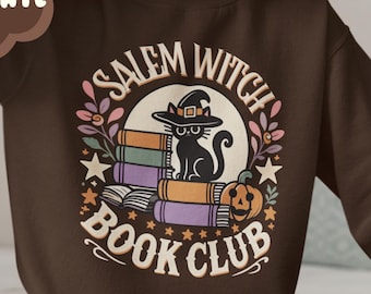 Könnte beinhalten: Ein braunes Sweatshirt mit dem Text "Salem Witch Book Club" in einem Kreis, auf dem ein schwarzer Kater mit einer Hexenmütze auf einem Stapel Bücher sitzt. Der Kater schaut nach links. Um den Kreis herum befinden sich Sterne und Blumen. Das Sweatshirt wird von einer Person getragen, die auf einem Bett sitzt.