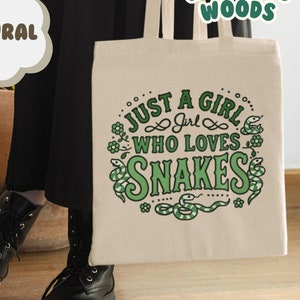 May include: A natural canvas tote bag with green text that reads "Just a girl who loves snakes". The text is surrounded by a green snake design. The bag is being held by a person wearing black boots.