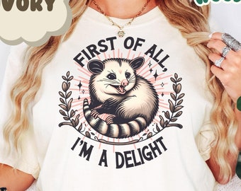 May include: A white t-shirt with a graphic of a possum with the text "First of all, I'm a delight." The shirt is a light ivory color.