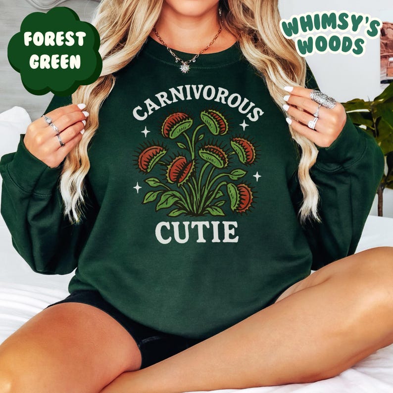 May include: Forest green sweatshirt with a graphic of a Venus flytrap plant. The text "CARNIVOROUS CUTIE" is printed in white. The sweatshirt is a solid forest green color.