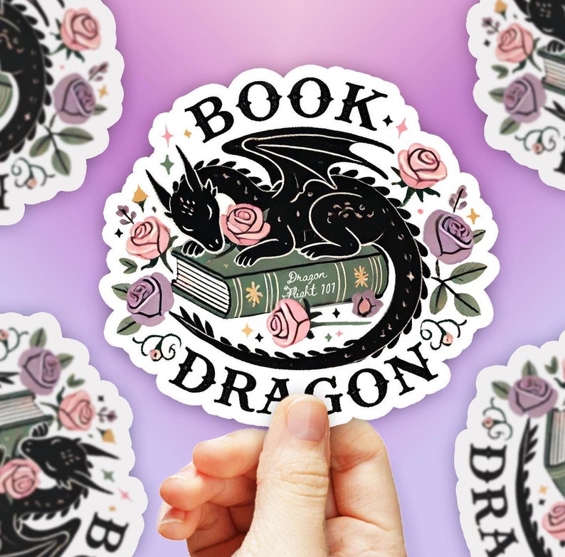 May include: A white sticker with a black dragon curled up asleep on a green book. The book has the words "Dragon Flight 101" on the cover. The sticker is surrounded by pink and purple flowers and the words "Book Dragon" are written in a black, stylized font.