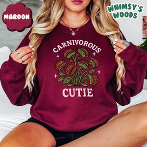 May include: Maroon sweatshirt with a graphic of a Venus flytrap plant and the words "CARNIVOROUS CUTIE" in white. The sweatshirt also has the text "WHIMSY'S WOODS" in green. The design features green leaves and orange and green flytraps.