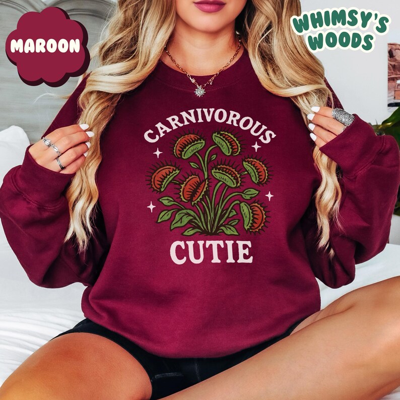 May include: Maroon sweatshirt with a graphic of a Venus flytrap plant and the words "CARNIVOROUS CUTIE" in white. The sweatshirt also has the text "WHIMSY'S WOODS" in green. The design features green leaves and orange and green flytraps.