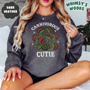 May include: Dark heather gray sweatshirt with a graphic of a Venus flytrap plant. The text "CARNIVOROUS CUTIE" is printed in white above and below the plant illustration. The words "DARK HEATHER" and "WHIMSY'S WOODS" are also visible.