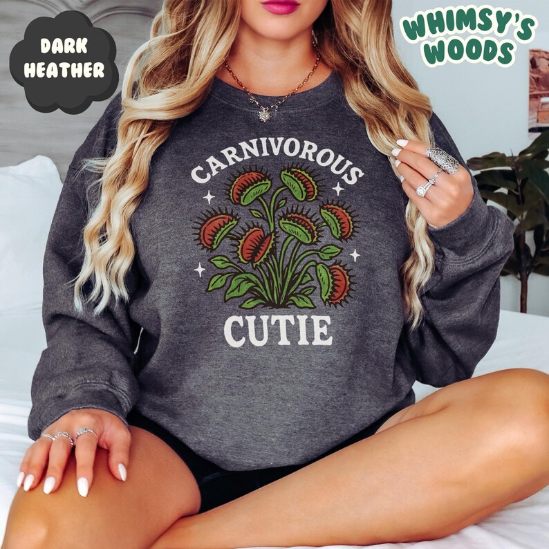 May include: Dark heather gray sweatshirt with a graphic of a Venus flytrap plant. The text "CARNIVOROUS CUTIE" is printed in white above and below the plant illustration. The words "DARK HEATHER" and "WHIMSY'S WOODS" are also visible.