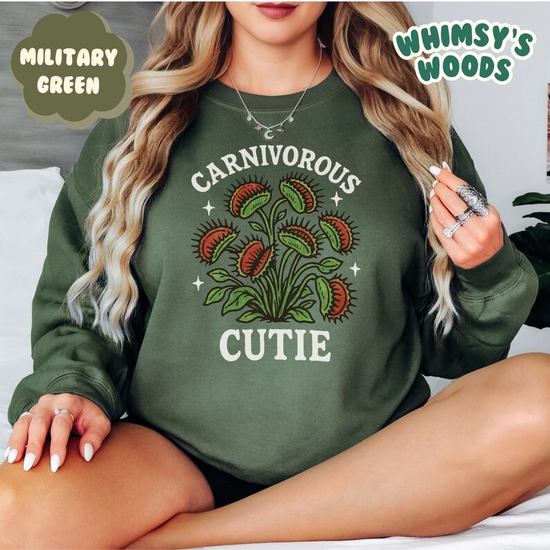 May include: Military green sweatshirt with a graphic of a Venus flytrap plant. The text "CARNIVOROUS CUTIE" is printed below the plant. The sweatshirt is a solid color and has long sleeves.