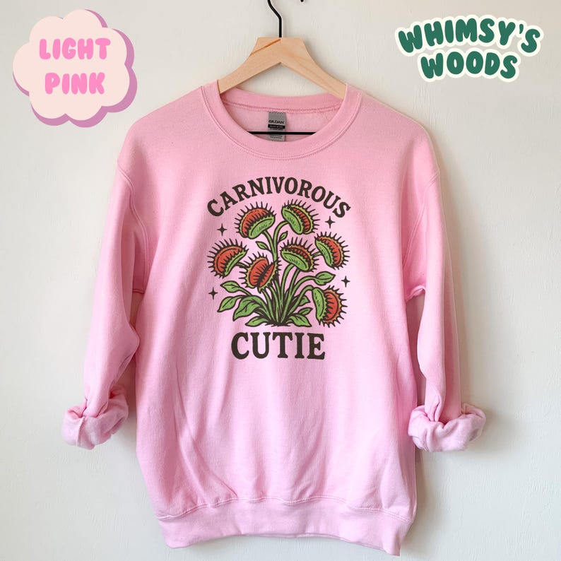 May include: Light pink sweatshirt with a graphic of a Venus flytrap plant. The text "CARNIVOROUS CUTIE" is printed in black above and below the plant illustration. The words "LIGHT PINK" and "WHIMSY'S WOODS" are also in the image.