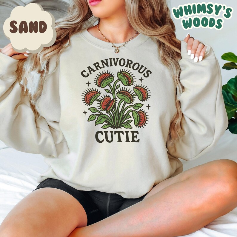 May include: Sand-colored sweatshirt featuring a graphic of a Venus flytrap with the words "CARNIVOROUS CUTIE". The design includes green leaves and red traps. The sweatshirt has a crew neck and long sleeves.