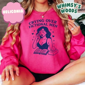 K&ouml;nnte beinhalten: Ein pinkfarbenes Sweatshirt mit dem Text "Crying Over Fictional Men" und einer Grafik einer weinenden Frau. Die Frau tr&auml;gt einen dunkelblauen BH und sitzt vor einem offenen Buch und einer Schachtel Taschent&uuml;cher. Das Sweatshirt hat lange &Auml;rmel.
