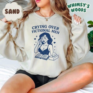 K&ouml;nnte beinhalten: Sandfarbenes Sweatshirt mit dem Text "Crying Over Fictional Men" und einer blauen Illustration einer weinenden Frau. Die Grafik enth&auml;lt ein offenes Buch, Taschent&uuml;cher und Sterne. Das Sweatshirt hat lange &Auml;rmel.
