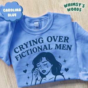 K&ouml;nnte beinhalten: Hellblaues Sweatshirt mit dem Text "Crying Over Fictional Men" und einer Illustration einer weinenden Frau. Das Design enth&auml;lt Sterne und Herzen. Der Markenname "Whimsy's Woods" ist oben rechts zu sehen.