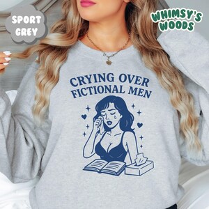 K&ouml;nnte beinhalten: Sportgraues Sweatshirt mit dem Text "Crying Over Fictional Men" und einer blauen Illustration einer weinenden Frau. Die Illustration zeigt ein Buch, Taschent&uuml;cher, Herzen und Sterne. Von Whimsy's Woods.