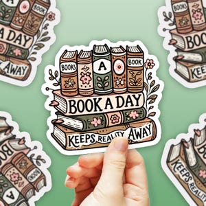 以下が含まれることがあります： 「Book a Day Keeps Reality Away」というテキストが書かれた本の積み重ねのステッカー。本は花で飾られており、テキストは茶色とピンクです。