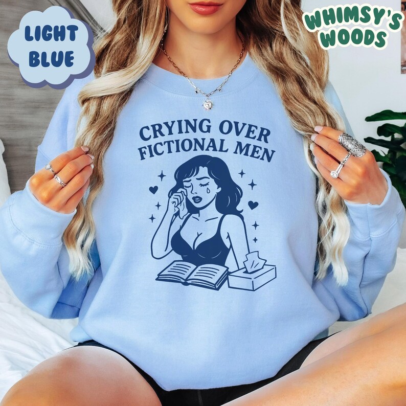 K&ouml;nnte beinhalten: Hellblaues Sweatshirt mit dem Text "Crying Over Fictional Men" und einer Grafik einer weinenden Frau. Das Sweatshirt hat auch den Text "Whimsy's Woods" in der oberen rechten Ecke.