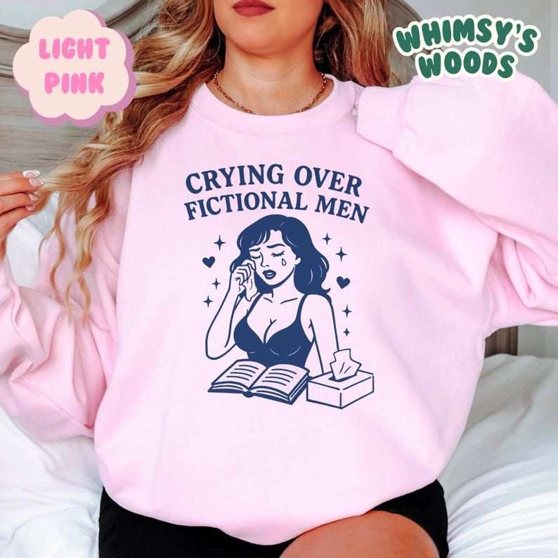 K&ouml;nnte beinhalten: Hellrosa Sweatshirt mit dem Text "Crying Over Fictional Men" &uuml;ber einer Grafik einer weinenden Frau. Die Grafik enth&auml;lt ein Buch, Taschent&uuml;cher und Herzen. Der Markenname "Whimsy's Woods" ist in gr&uuml;ner Schrift.