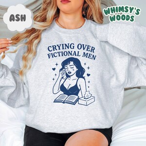 K&ouml;nnte beinhalten: Hellgraues Sweatshirt mit dem Text "Crying Over Fictional Men" und einer Grafik einer weinenden Frau. Das Design umfasst Herzen, Sterne, ein Buch und Taschent&uuml;cher. Der Markenname "Whimsy's Woods" ist ebenfalls sichtbar.