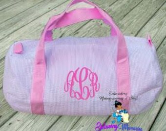 May include: A lavender and white striped duffel bag with pink handles and a pink monogram. The bag has a rectangular shape and is made of a seersucker fabric. The bag is designed for travel or carrying items.