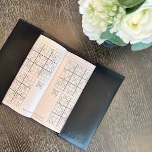 May include: An open black leather-bound book with a Sudoku puzzle on each page. The book is open to page 112 and 114. The text "VIA VITA" is printed on the cover.